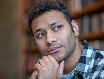 Young adult man with chin on hid hands staring into distance in deep thought